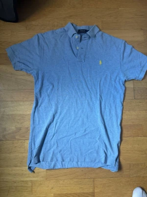 Blå pikétröja från Polo Ralph Lauren - Klassisk blå pikétröja från Polo Ralph Lauren med korta ärmar och krage. Tröjan har knappar vid halsen och den ikoniska gula logotypen broderad på bröstet. Tillverkad i mjuk bomull för en bekväm känsla.