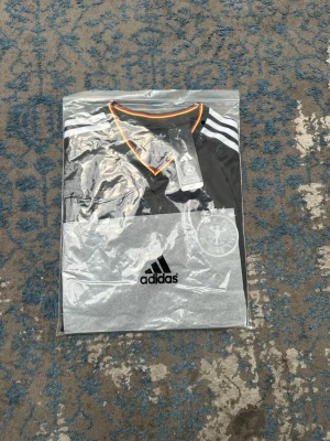 Svart Adidas Tyskland matchtröja - Säljer en svart Tyskland matchtröja från Adidas med vita ränder på axlarna och orange detaljer vid krage och ärmslut. Tröjan har Tysklands landslagsmärke på bröstet och Adidas-logga framtill. Perfekt för fotbollsfans som vill sticka ut på planen.