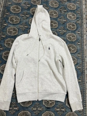 Ljusgrå hoodie från Polo Ralph Lauren - Snygg ljusgrå hoodie med dragkedja från Polo Ralph Lauren. Klassisk design med luva och dragsko, två fickor framtill och ikoniska broderade loggan på bröstet. Perfekt för en chill och stilren look.