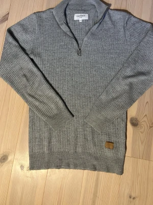 Lindebergh stickad quarterzip - Snygg grå stickad tröja från Lindbergh med ribbad struktur och halvzip i halsen. Tröjan har långa ärmar, hög krage och en liten brun patch nertill. Tunn och go stickad tröja perfekt nu till sommarkvällar