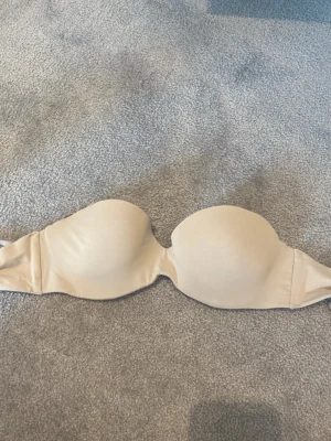 Beige axelbandslös bh - Säljer en beige axelbandslös bh med släta kupor och avtagbara axelband. Bh:n har en enkel och stilren design, perfekt under ljusa toppar. Materialet känns mjukt och stretchigt för extra komfort.