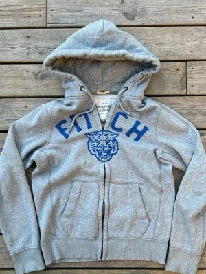 Grå hoodie från Abercrombie & Fitch - Säljer en grå zip-hoodie från Abercrombie & Fitch med blå broderad text 'FITCH' och ett coolt tigerhuvud på bröstet. Hoodien är riktigt fet o passar för lite kyligare dagar under sommaren. Hör av dig vid frågor 