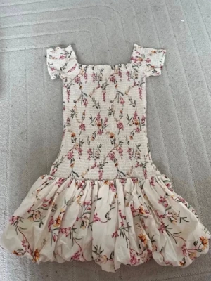 Blommig kortklänning med volang - Supersöt kortklänning med blommigt mönster i rosa, gul och grön på vit botten. Klänningen har puffiga korta ärmar, smockad överdel och volangig kjol. Perfekt för varma dagar och ger en romantisk vibe.
