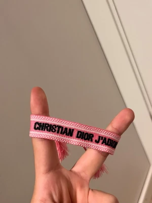 Christian Dior J'Adior rosa armband - Säljer ett rosa tygarmband från Christian Dior med texten 'J'Adior' i svart. Armbandet har vävd struktur och tofsar i ändarna, perfekt för att ge din outfit en trendig touch.