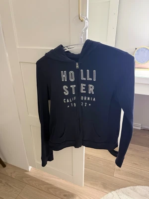 Svart hoodie från Hollister - Snygg svart hoodie från Hollister med dragkedja framtill och vit text på bröstet där det står 'Hollister California 1922'. Hoodien har två fickor fram och en klassisk huva. Perfekt för en avslappnad och cool stil.