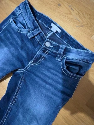 Blå jeans med broderade bakfickor 💙 - Säljer ett par blå jeans med snygga slitningar och kontrastsömmar. Bakfickorna har stora broderade mönster i vitt och blått som verkligen sticker ut. Priset kan diskuteras! Klassisk femficksmodell med knappgylf och bälteshällor. Perfekta för dig som vill ha något unikt!