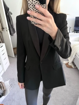 Svart kavaj med satinkrage - Stilren svart kavaj med smal passform och elegant satinkrage. Kavajen har två fickor med lock framtill och knäppning med en knapp. Perfekt för en dressad look och funkar till många olika outfits. Storlek Small och aldrig använd! Nypris: Ca 900 kr