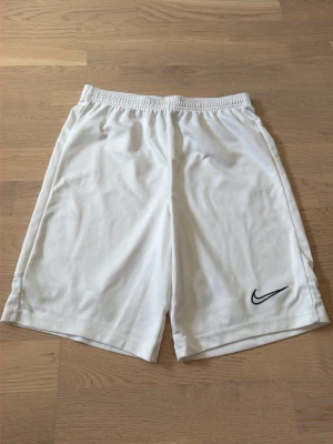 Vita sportshorts från Nike - Snygga vita sportshorts från Nike med klassisk svart logga på ena benet. Tillverkade i ett lätt och svalt polyestermaterial som passar perfekt för träning eller sportaktiviteter. Enkel och stilren design med normal passform.