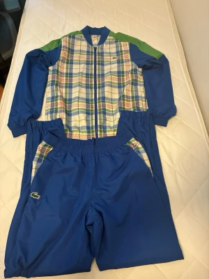 Blått träningsset från Lacoste Sport - Snyggt träningsset från Lacoste Sport med blå byxor och jacka. Jackan har rutigt mönster i blått, grönt, rött och vitt framtill samt gröna detaljer på axlarna. Byxorna har matchande rutiga detaljer vid fickorna och klassisk Lacoste-logga. Perfekt för sport eller chill.