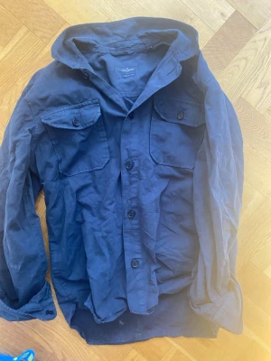 Mörkblå overshirt - Säljer en mörkblå skjorta med två bröstfickor och knappar framtill. Skjortan har lång ärm och klassisk krage, perfekt för en avslappnad men ändå stilren look. 