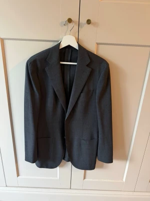 Traveller kavaj från Suitsupply grå - Stilren mörkgrå kavaj från Suitsupply, modell Traveller. Klassisk passform med två framfickor och en bröstficka. Perfekt för dig som vill ha en tidlös look. Tillverkad i ett slätt tyg som ger en elegant känsla. Nypris 4500
