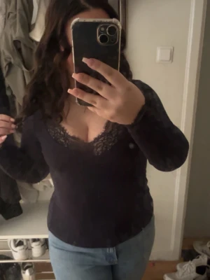 Svart långärmad topp med spets - Snygg svart långärmad topp med v-ringning och spetsdetaljer vid halsringningen. Figurnära passform som framhäver siluetten. Perfekt att styla med jeans eller kjol för en trendig look.
