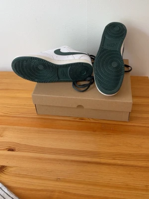 Nike vita och gröna sneakers - Säljer ett par vita Nike sneakers med gröna detaljer och sula. Klassisk låg modell med perforerad tåbox och grönt Nike-märke på hälen. Skorna har vita ovandelar i läder och gröna snören. Perfekta för dig som gillar stilrena och sportiga sneakers.