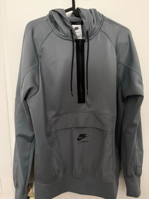 Grå Nike Air Max hoodie med dragkedja - Snygg grå hoodie från Nike Air Max med huva och svarta dragsnören. Tröjan har en halv dragkedja framtill och en stor magficka med Nike Air Max-logga. Tillverkad i mjukt och värmande material, perfekt för chill eller träning.