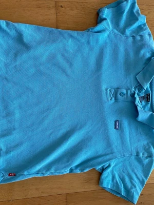 Blå piké från Levi's - Snygg ljusblå pikétröja från Levi's med klassisk krage och knappar framtill. Tillverkad i bomull för en skön känsla. Perfekt till jeans eller shorts för en avslappnad stil. Levi's-logga på bröstet och röd etikett i sidan.