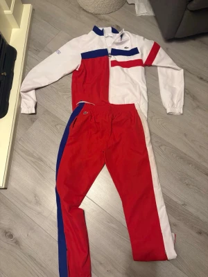 Lacoste Sport tracksuit röd/vit/blå - Säljer ett retroinspirerat tracksuit-set från Lacoste Sport i färgerna röd, vit och blå. Jackan har dragkedja, ståkrage och färgblocksmönster, medan byxorna har resår i midjan och matchande färgpaneler längs sidorna. Perfekt för dig som gillar sportig och stilren look.