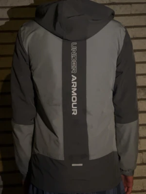 Grå vindjacka från Under Armour - Snygg grå vindjacka från Under Armour med huva och dragkedja framtill. Jackan har två nyanser av grått, logga på bröstet och stort vertikalt tryck på ryggen. Perfekt för blåsiga dagar och sportiga outfits.