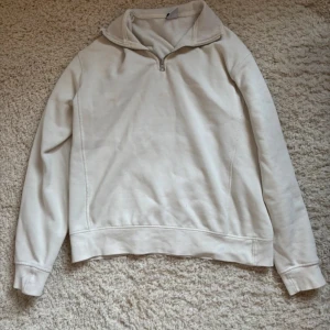 Vit halvzip hoodie - Säljer en vit hoodie med halv dragkedja och hög krage. Hoodien har långa ärmar och ribbade muddar vid ärmslut och nederkant. Perfekt för en avslappnad och stilren look. Materialet känns mjukt och bekvämt mot huden.