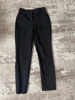 Svarta raka kostymbyxor - Stilrena svarta kostymbyxor med raka ben och pressveck. Byxorna har hällor för skärp och sidofickor. Perfekta för en clean och klassisk look. Materialet ser ut att vara en mjuk och lite stretchig mix, vilket ger en bekväm passform.