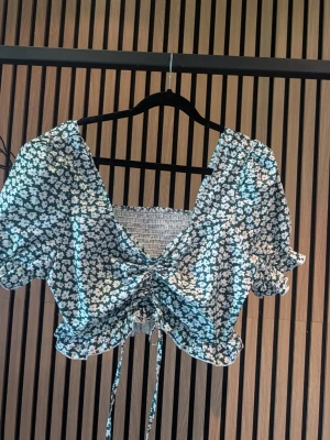 Blommig croppad blus med snörning - Supersöt blommig croppad blus i grönt och vitt med puffärmar och snörning framtill. Toppen har v-ringning, volangkant och elastisk rygg för skön passform. Perfekt för sommarens alla tillfällen! Storlek L men är liten i storleken, den är mer en M
