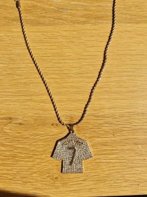 Guldhalsband med tröjhänge och stenar - Snyggt halsband i guldfärg med ett hänge format som en ronaldotröja som är , täckt av glittrande stenar och siffran 7 i mitten. Kedjan är tvinnad och har ett justerbart lås. Perfekt för dig som gillar sportig stil och bling. själva bandet och tröjan är gjord av silver som det stog när jag köpte den för 600 kr så stog det att den var gjord av silver och täckt i guld färgat stål så att jag säljer den för halva priset