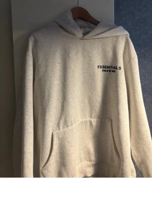 Grå Essentials Fear of God hoodie - Snygg ljusgrå hoodie från Fear of God Essentials med svart logga tryckt på bröstet och ryggen. Klassisk känguruficka framtill och stor huva. Perfekt för dig som gillar streetwear och vill ha en stilren look.