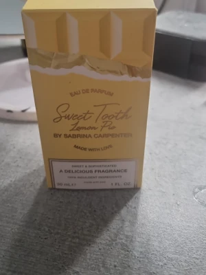 Sweet Tooth Lemon Pie parfym - Söt och fräsch parfym i doften Sweet Tooth Lemon Pie från Sabrina Carpenter. Förpackningen är gul med ett bakverksinspirerat mönster och text i guld. Innehåller 30 ml Eau de Parfum och har en lekfull, modern design som sticker ut.