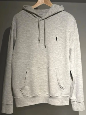 Ljusgrå hoodie från Polo Ralph Lauren - Snygg ljusgrå hoodie från Polo Ralph Lauren med klassisk huva och dragsnören. Har en stor magficka och den ikoniska broderade loggan i svart på bröstet. Tillverkad i mjukt bomullsmaterial som känns riktigt skönt mot huden.
