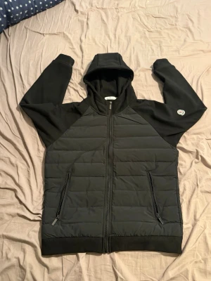 Svart quiltad hoodiejacka - Svart hoodiejacka med quiltade paneler framtill och baktill, dragkedja hela vägen och två fickor med dragkedja. Jackan har huva och ribbade muddar. Liten patch på ärmen för extra detalj. QR koden fungerar