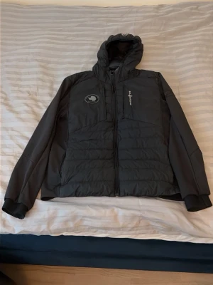 Svart pufferjacka från Peak Performance - Snygg svart pufferjacka från Peak Performance med huva och dragkedja framtill. Jackan har en bröstficka med logga, två sidofickor och diskreta detaljer. Perfekt för kalla dagar och riktigt skön passform. Klassisk design med sportig touch.
