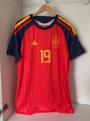 Spaniens matchtröja Lamine Yamal #19 - Säljer en officiell röd Spanien fotbollströja från Adidas med gula detaljer, nummer 19 och Lamine Yamal på ryggen. Tröjan har blå ärmar med gula ränder på axlarna och är tillverkad i ett lätt, ventilerande material perfekt för träning eller match.