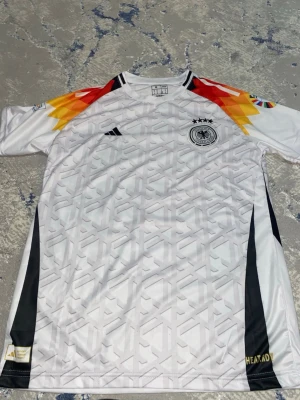 Tyskland fotbollströja - Säljer en vit T-shirt från Adidas med Tysklands landslagsemblem och fyra stjärnor på bröstet. T-shirten har färgglada detaljer i rött, gult och svart på axlarna samt diskret mönster över hela framsidan. Tillverkad i polyester. HAR INTE ANVÄNT OCH KAN SÄNKA PRIS