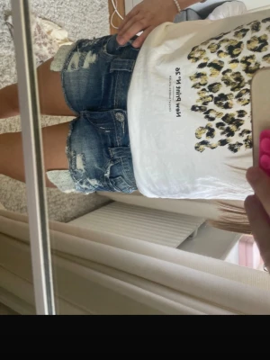 Blå slitna jeansshorts med detaljer - Säljer ett par blå jeansshorts med slitna detaljer och råa kanter. Shortsen har klassisk femficksdesign och en cool, avslappnad look. Perfekta för varma dagar och enkel att matcha med olika toppar.