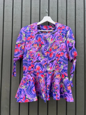Vintage blommig peplumblus - Färgstark blus med peplum och lång ärm i ett virvlande blommönster i lila, rött, grönt och rosa. Blusen har knappar framtill och knytband vid ärmsluten. 