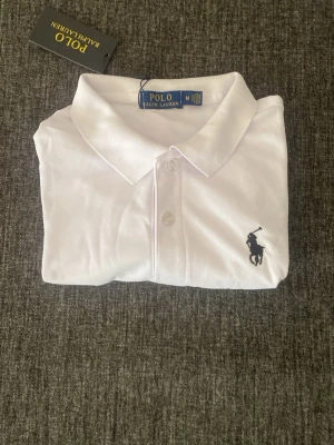Vit skojets från Polo Ralph Lauren - Klassisk vit pikétröja från Polo Ralph Lauren med korta ärmar och krage. Tröjan har knappar framtill och den ikoniska broderade loggan i svart på bröstet. Perfekt till jeans eller shorts för en clean och stilren look.