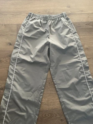 Grå jogging byxor  med vita detaljer - Säljer ett par grå joggingbyxor med raka ben och vita piping längs sidorna. Byxorna har resår i midjan för skön passform och är tillverkade i ett mjukt, lätt material. Perfekta för en avslappnad och trendig look.