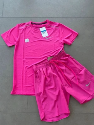 Rosa träningsset från Nike - Snyggt rosa träningsset från Nike med t-shirt och shorts. T-shirten har rund hals och korta ärmar, medan shortsen har resårmidja och dragsko för skön passform. Båda plaggen har Nike-logga framtill och är tillverkade i lätt, snabbtorkande material.