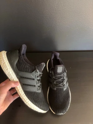 Svarta Adidas Ultraboost sneakers - Säljer ett par svarta Adidas Ultraboost sneakers med stickad ovandel och klassiska tre ränder på sidan. Skorna har en vit, responsiv Boost-sula och svart hälkappa. Perfekta för dig som gillar sportig och stilren look. Priset är ej hugget i sten och jag skickar inom en dag! Hör av dig vid minsta frågor!
