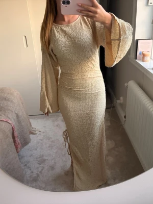 Beige långklänning med vida ärmar från NA-KD - Trendig beige långklänning med strukturerat tyg och vida, långa ärmar. Klänningen har en figurnära passform och lätt rynkade detaljer i sidan. Ordenarie pris 800kr och har prislappen på.