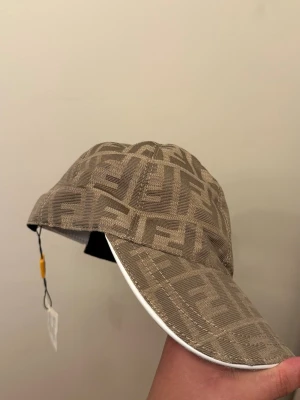 Beige Fendi keps - Säljer en beige keps från Fendi med det klassiska FF-monogrammet över hela kepsen. Kepsen har böjd skärm med vit kant och justerbart spänne bak i vitt läder. Materialet är tyg med exklusiv känsla och kepsen har originaltaggar kvar.