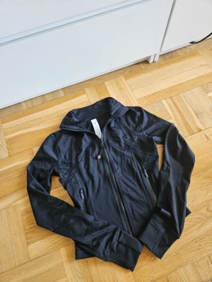 Lululemon Define Jacket Nulu - Sportig svart jacka från Lululemon Nulu Define Jacket med dragkedja framtill och två fickor med dragkedja. Jackan har en figurnära passform, hög krage och långa ärmar. Perfekt för träning eller som ett extra lager till vardags. Nypris 1380kr