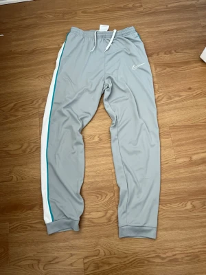 Grå Nike träningsbyxor med vit rand - Snygga grå träningsbyxor från Nike med vit och turkos rand längs sidan. Byxorna har resår i midjan med vit dragsko och muddar vid bensluten. Tillverkade i ett mjukt och stretchigt material som passar perfekt till träning eller chill.