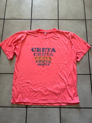 Korallfärgad t-shirt med CRETA-tryck storlek XL/XXL - Säljer en korallfärgad t-shirt med trycket 'CRETA' i olika färger och 'GREECE' på bröstet. T-shirten har rund hals och korta ärmar, perfekt för dig som gillar färg och vill sticka ut lite extra. Materialet känns mjukt och skönt mot huden.