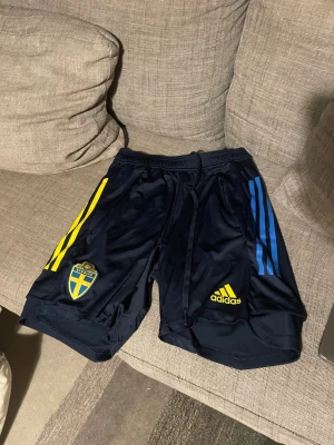 Blå Adidas fotbollsshorts Sverige - Mörkblå fotbollsshorts från Adidas med Sveriges landslagsemblem på vänster ben och gula samt blå ränder längs sidorna. Shortsen har snörning i midjan och är tillverkade i ett lätt, syntetiskt material som passar perfekt för träning och VM 🇸🇪🇸🇪‼️‼️