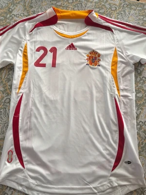 Spanien VM 2006 bortatröja, David Villa 21 - Säljer en vit Spanien fotbollströja från Adidas med röd och gul detaljering, nummer 21 och David Villa på ryggen. Tröjan har landslagets emblem på bröstet och klassiska Adidas-ränder på axlarna. Perfekt för dig som älskar fotboll och vill stötta Spanien.