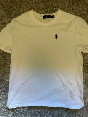 Vit t-shirt från Polo Ralph Lauren - Klassisk vit t-shirt från Polo Ralph Lauren med korta ärmar och rund hals. T-shirten har den ikoniska lilla marinblå polospelaren broderad på bröstet. Tillverkad i mjuk bomull för en skön känsla och enkel stil.