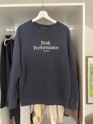 Mörkblå sweatshirt Peak Performance - Mörkblå sweatshirt från Peak Performance med vit logga tryckt på bröstet. Klassisk rund halsringning och ribbade muddar vid ärmslut och nederkant. Perfekt för en avslappnad och sportig stil.