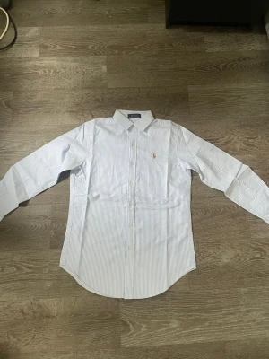 Blårandig skjorta från Ralph Lauren - Klassisk blårandig skjorta från Ralph Lauren med broderad logga på bröstet. Tillverkad i bomull för en skön och fräsch känsla. Perfekt för dig som gillar stilrena och fräscha plagg. Storlek M men skulle säga att den sitter som en S.
