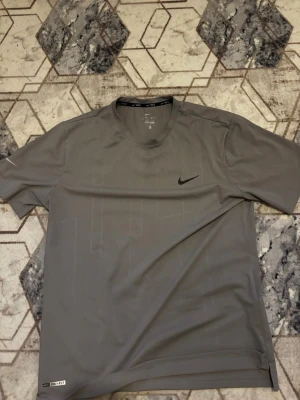 Grå Nike Dri-Fit träningströja - Snygg grå tränings-tshirt från Nike med Dri-Fit material som andas och transporterar bort svett. Klassisk rund halsringning och diskret Nike-logga på bröstet. Perfekt för gymmet eller löprundan.