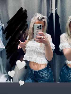 Vit spetsvolang topp - Söt croppad topp i vitt med volanger och spetsdetaljer. Ärmlös design och lätt transparent material som ger en romantisk vibe. Perfekt att styla med jeans eller kjol för en trendig look.